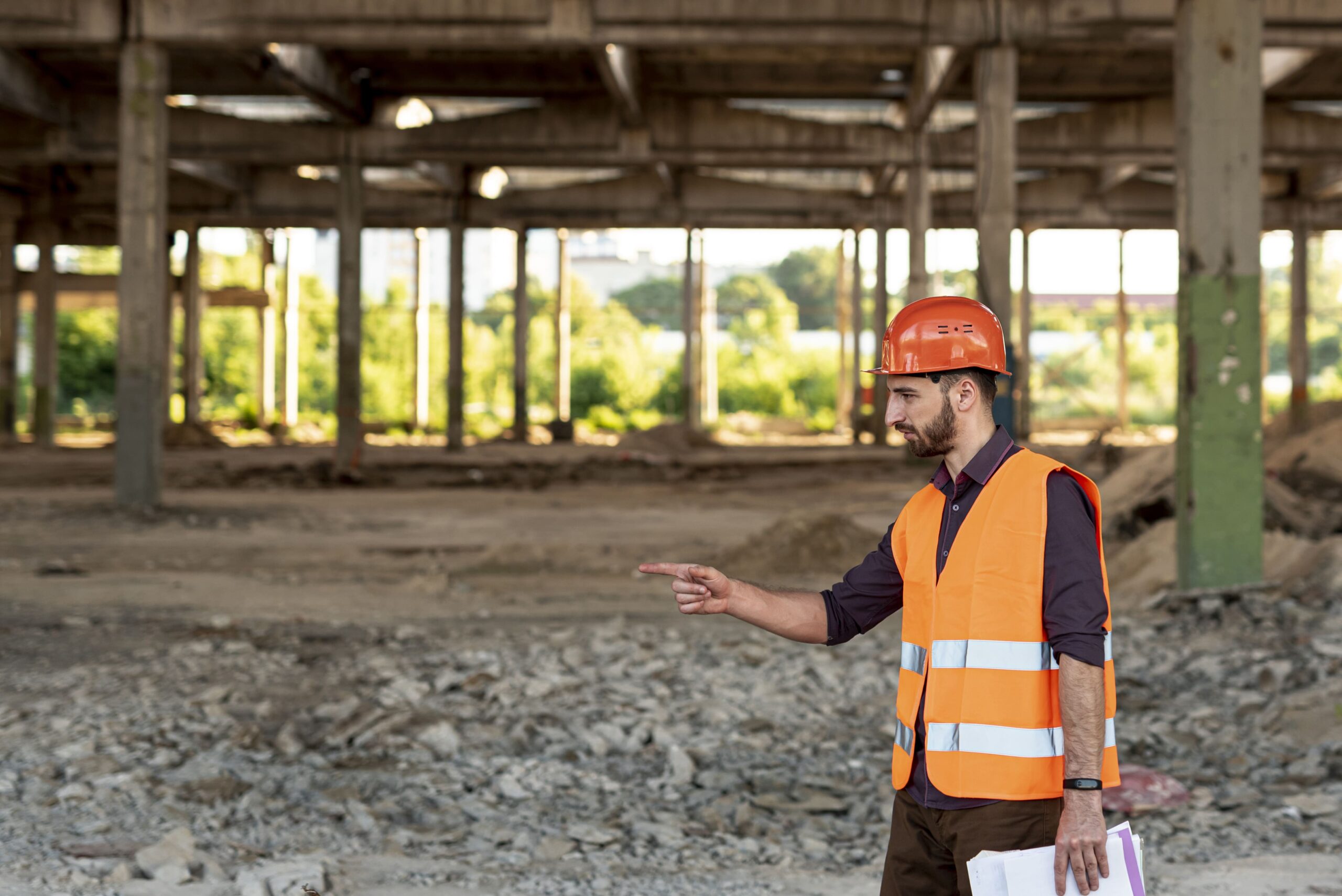 Construction site worker in orange safety vest and hard hat performing private utility locating field verification with GPR and electromagnetic methods near concrete columns.