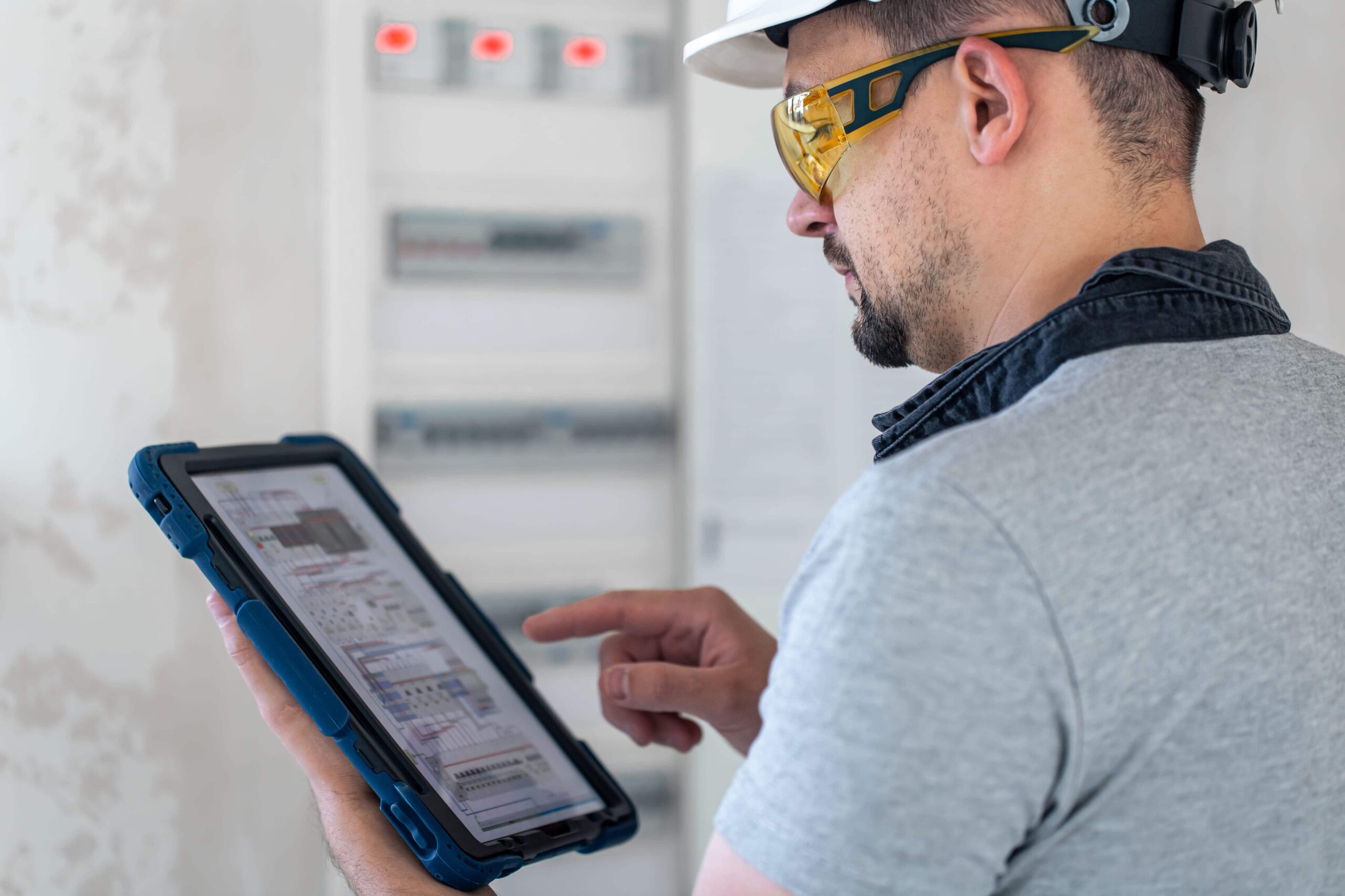 Construction worker in white hard hat and safety glasses using tablet near electrical panel for slab scanning before core drilling, saw cutting, and remodel planning.