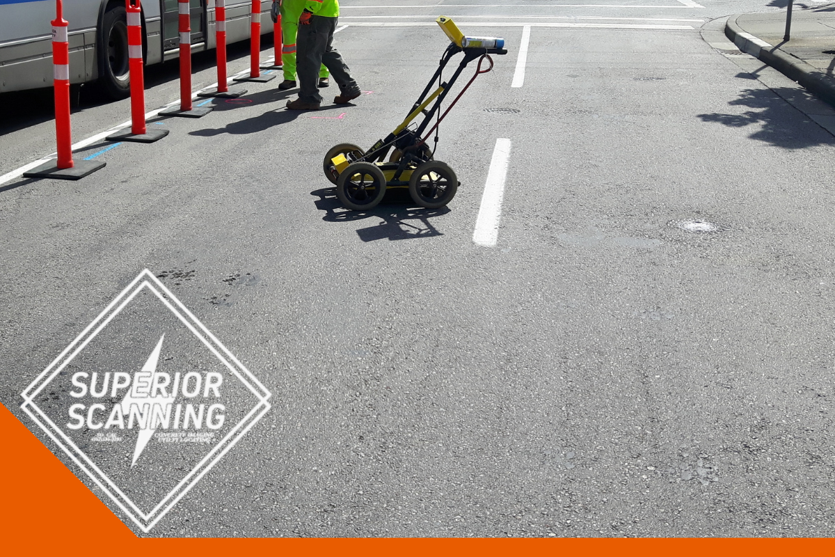 A ground penetrating radar cart is positioned on an asphalt road next to a bus and traffic cones.