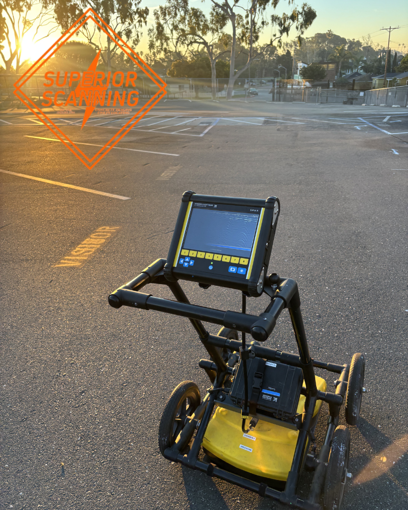 A ground penetrating radar cart with a display screen sits in a parking lot at sunset. Parking lines are visible on the asphalt and trees are in the background.
