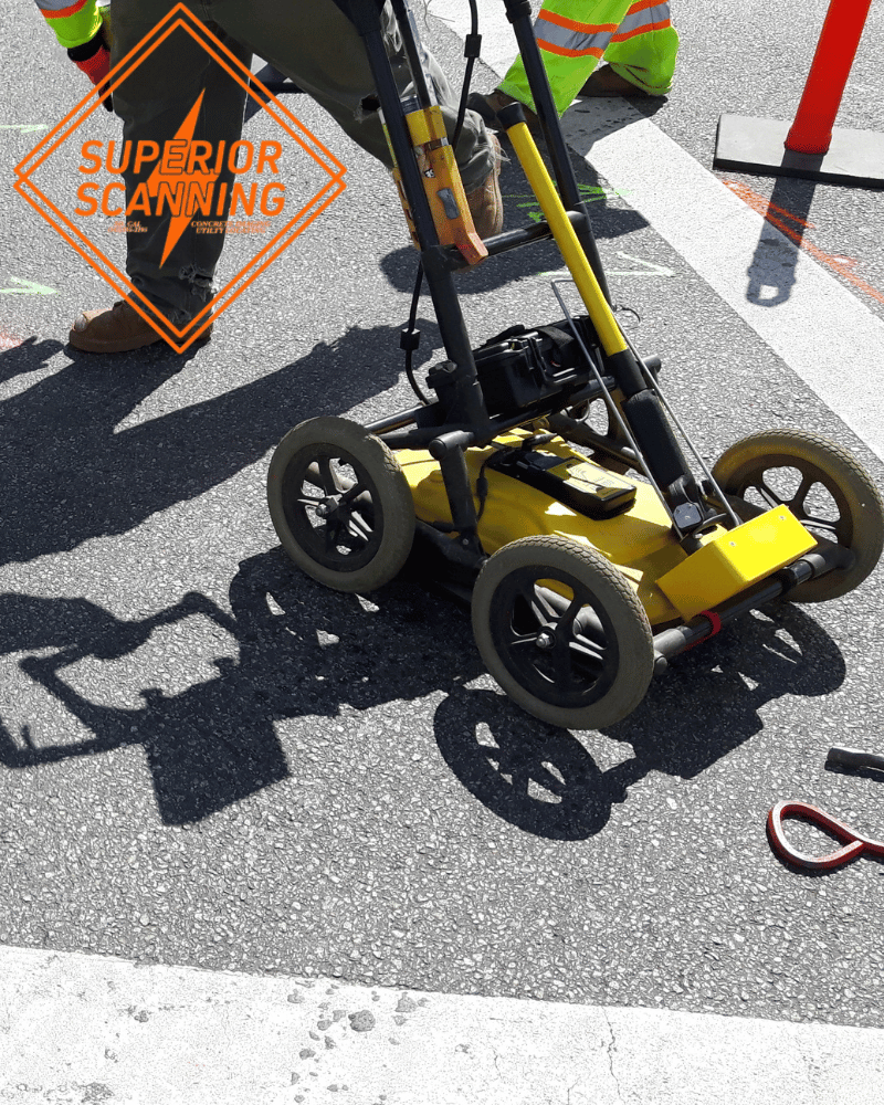 A yellow ground penetrating radar device with four wheels is on an asphalt surface, being operated by a person in high-visibility clothing.