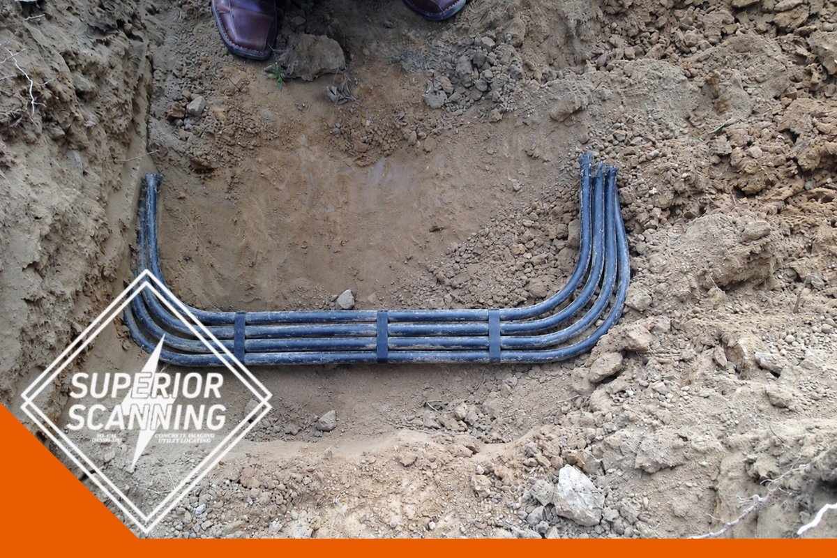 A U-shaped bundle of black pipes lies in a trench dug in the dirt. The pipes are held together by two dark blue straps.