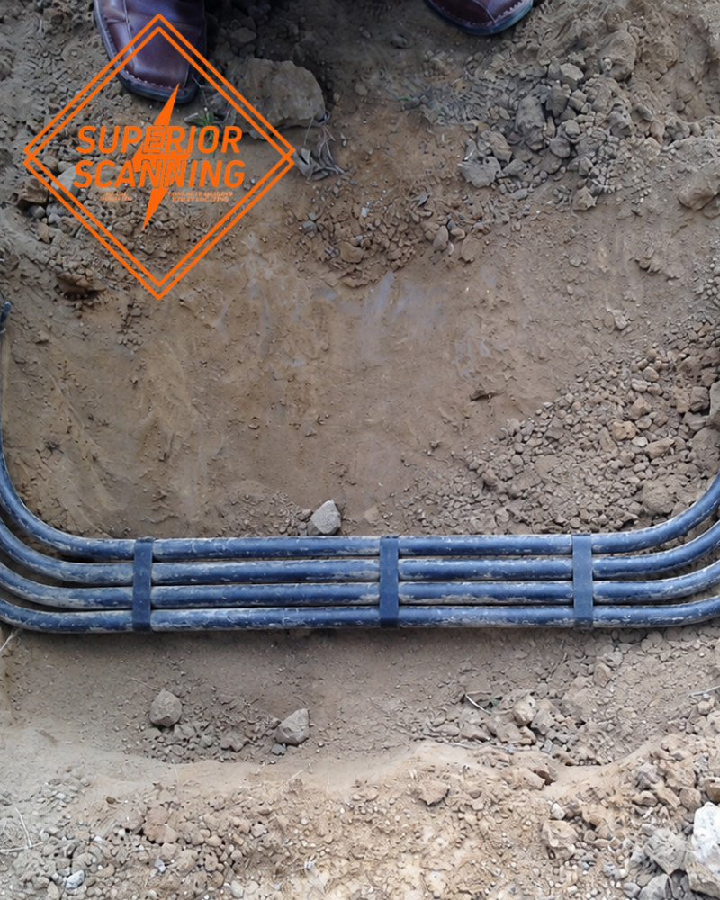Several dark, flexible pipes are bundled together with blue straps and laid in a trench of dirt.