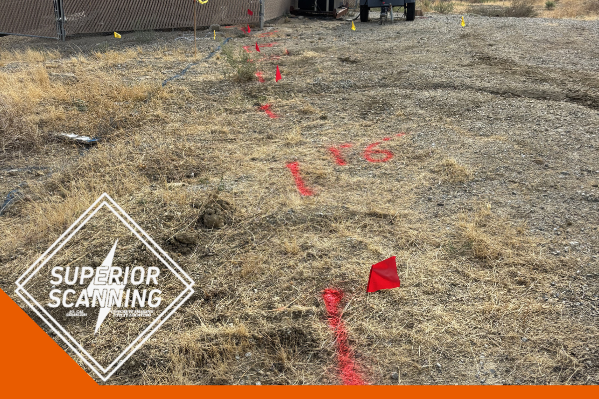 Red spray paint marks and small red flags indicate a line on dry, grassy ground. Yellow flags and a trailer are visible in the background near a fence.