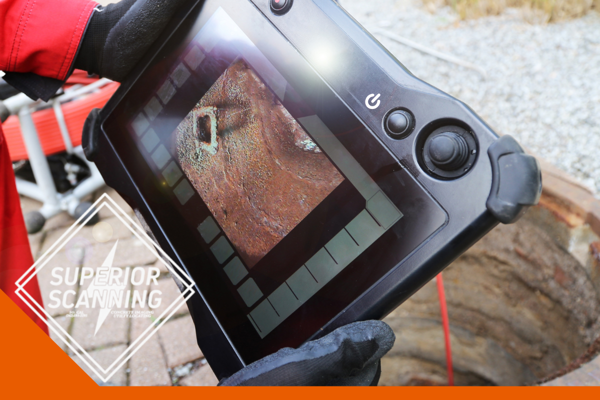 A person in red overalls holds a tablet displaying a camera feed of a sewer pipe, with the opening of the sewer visible below.