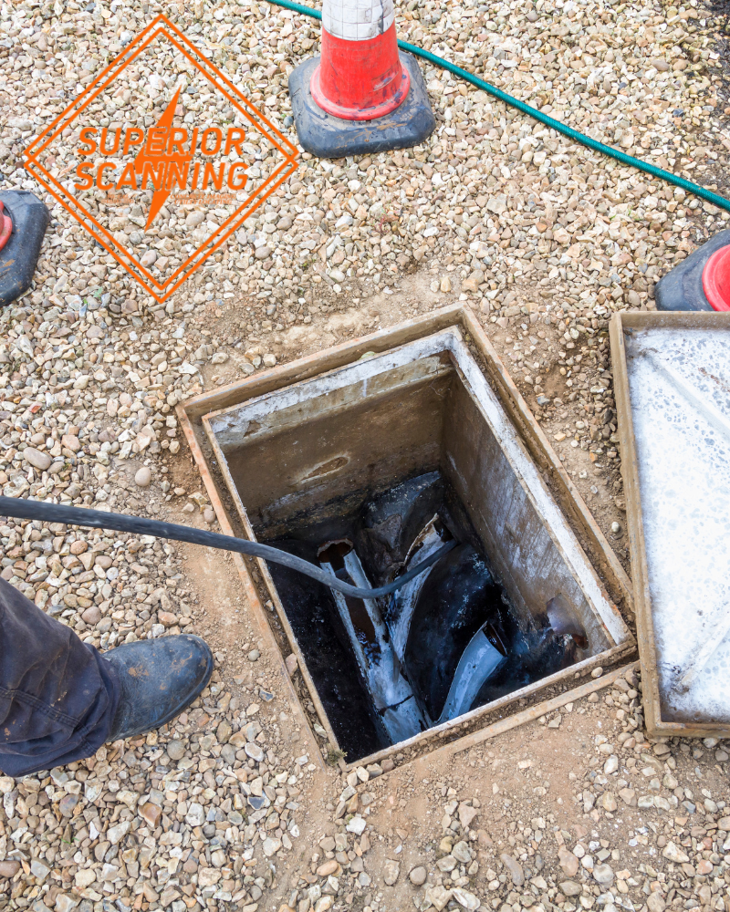 A person in boots is using a drain cleaning machine to clear a blocked drain. The drain cover is open, revealing debris inside. Orange traffic cones are visible in the background.