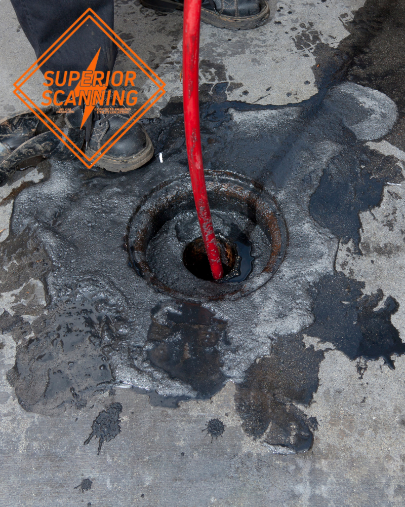 A red hose is inserted into a drain opening on a concrete surface, with dark liquid spilled around the drain.