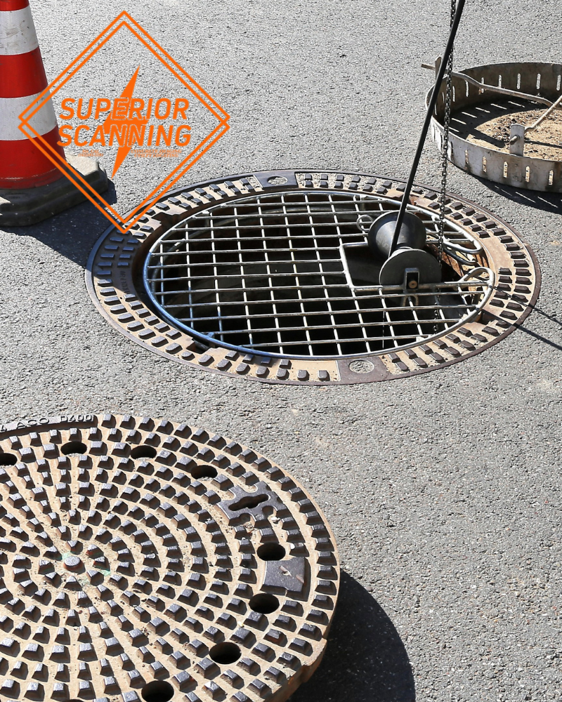 A sewer maintenance truck is parked on a street next to an open manhole with a camera on a pole lowered into it. A traffic cone is placed nearby.