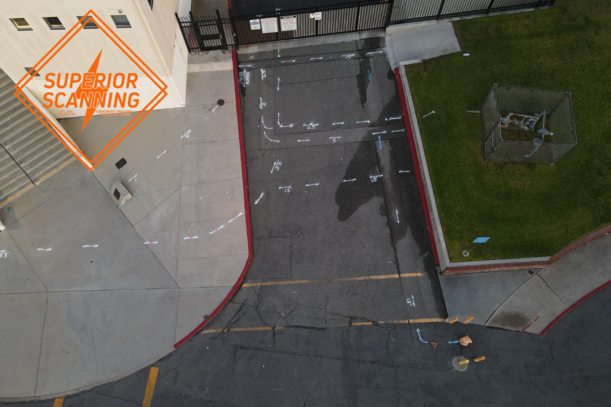 An aerial view of a schoolyard with painted directional arrows on the asphalt and a fenced enclosure on green area with equipment.