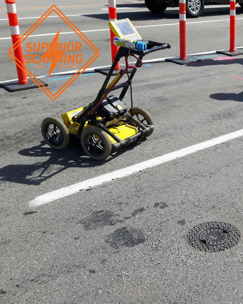 A yellow ground penetrating radar cart with a screen on top is parked on an asphalt road next to orange and white traffic cones.