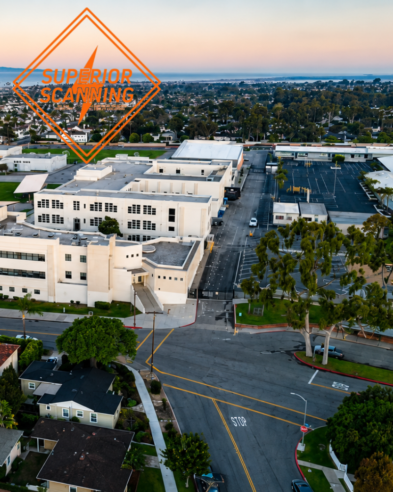 Aerial view of a large white building complex with a parking lot and surrounding residential neighborhood, with a distant ocean visible under a soft twilight sky.