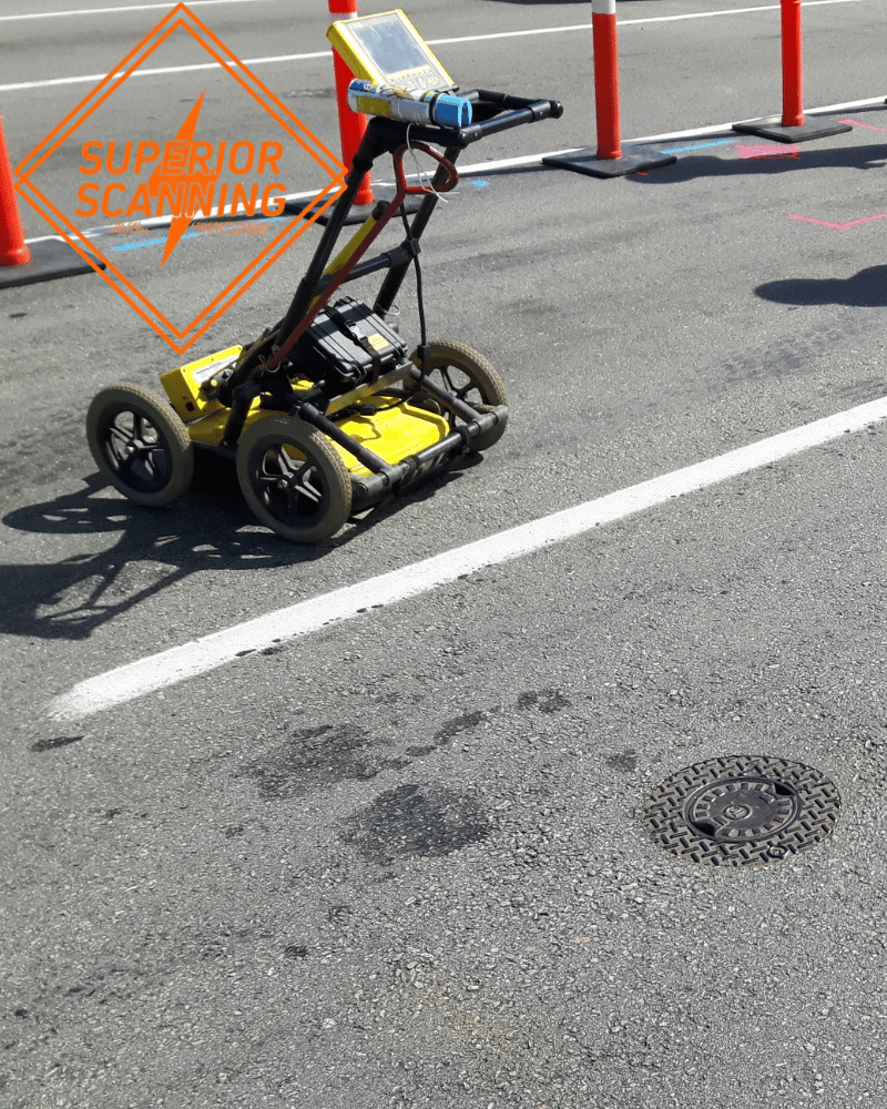 A yellow ground penetrating radar unit is on asphalt next to a manhole cover and orange traffic cones.