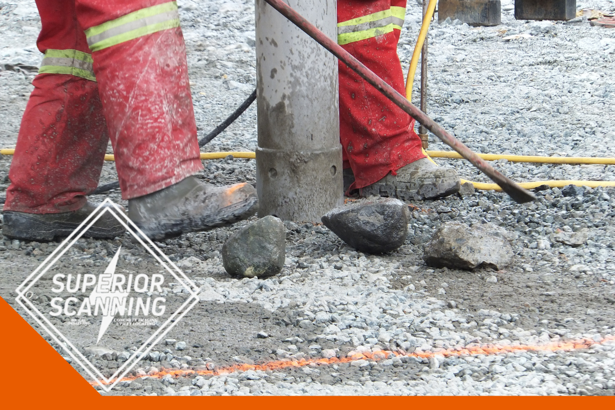 Workers in red overalls and muddy boots stand around a concrete pipe on a gravel surface. One worker holds a long tool, possibly a shovel or pry bar.