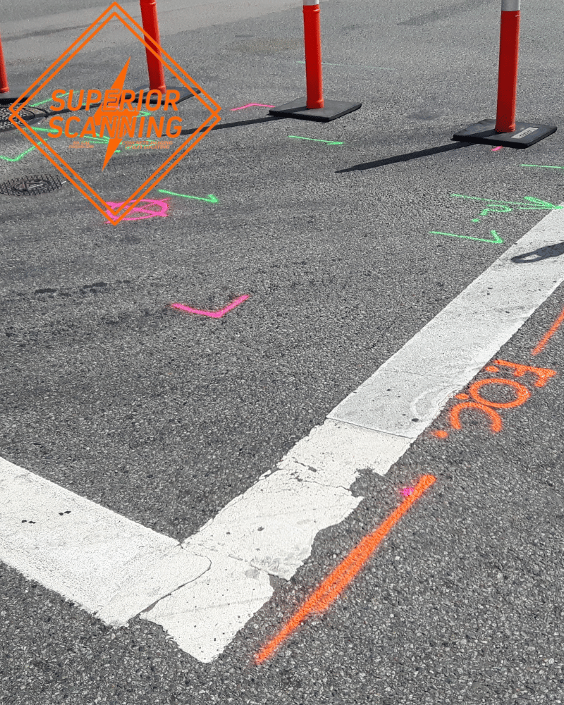 Orange and pink spray paint marks with arrows and lines are on an asphalt surface near a white painted line and orange traffic cones.