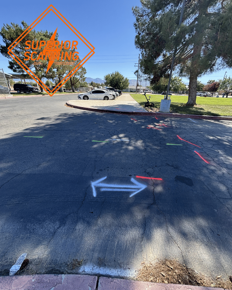 A white double-headed arrow is spray-painted on asphalt, with red and green spray-painted lines indicating directions in the background. White cars are parked in a lot behind the arrow.