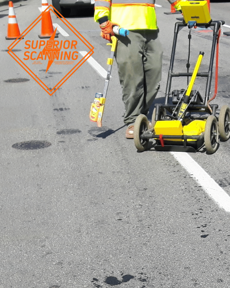 A person in a high-visibility vest uses a ground penetrating radar cart on a street, with traffic cones in the background.