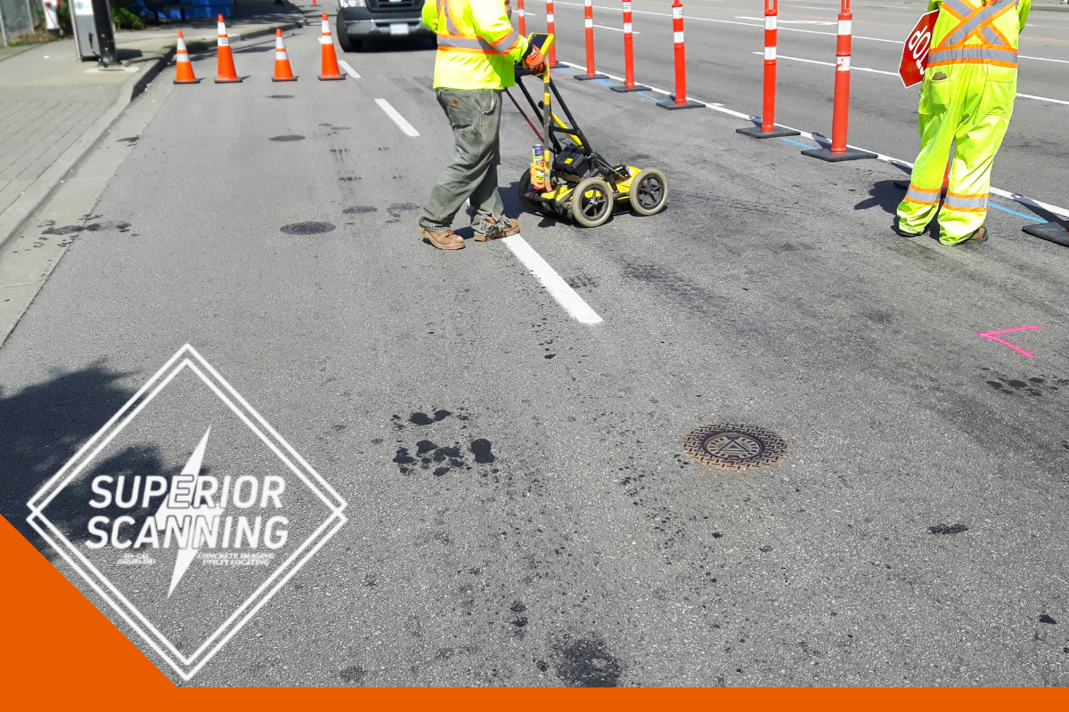 A person in a yellow vest uses a ground penetrating radar unit on a road, with traffic cones and safety markers visible.