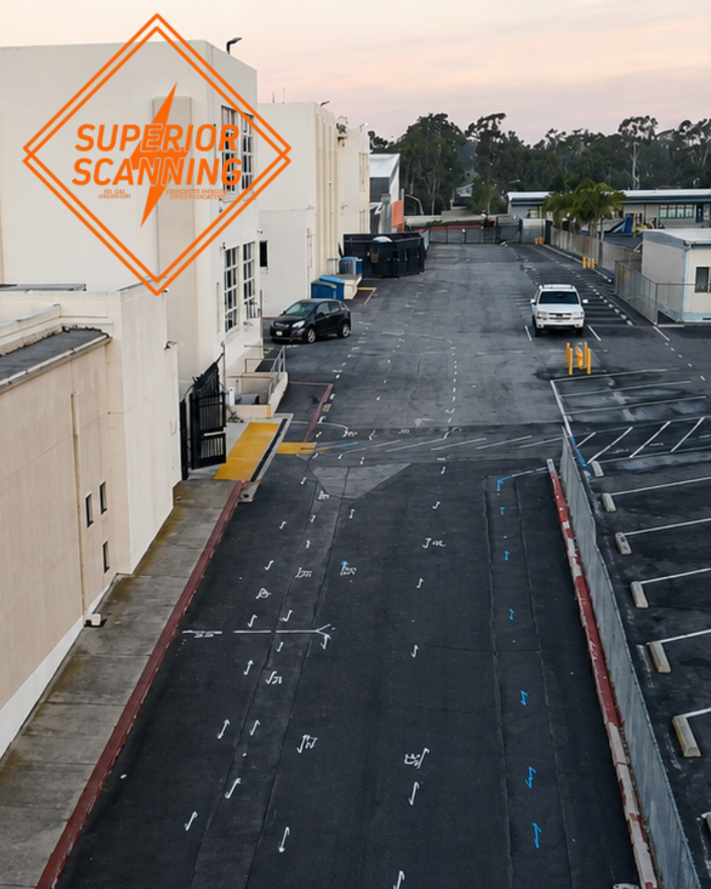 An aerial view of a paved road with parking spaces marked and a row of buildings on the left. Two cars are visible on the road, and trees line the horizon under a pale sky.